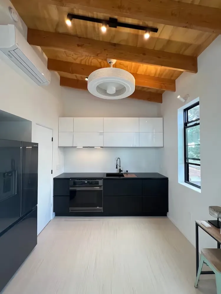 The Prince House ADU kitchen features minimalist black and white cabinetry with exposed wooden ceiling beams.