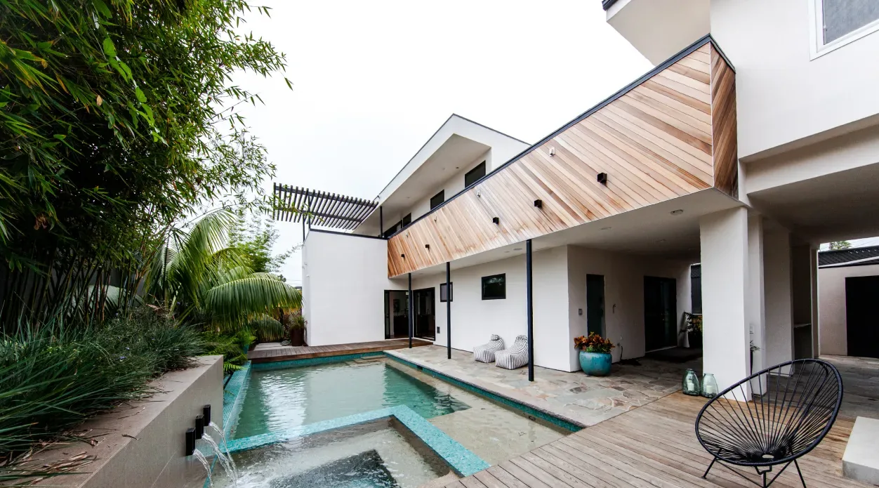 The Edinberg Residence features a pool adjacent to a covered patio with wood siding and a metal slat pergola above.