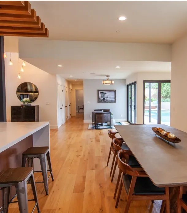 Interior view of an open dining and living space featuring wood floors and a concrete dining table facing large sliding glass doors.