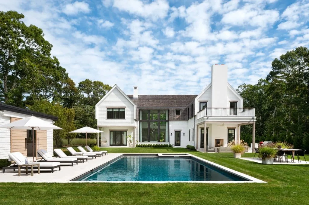 The Amagansett House features a symmetrical facade with gable roofs, double chimneys, and an outdoor pool area with lounge chairs and dining space.