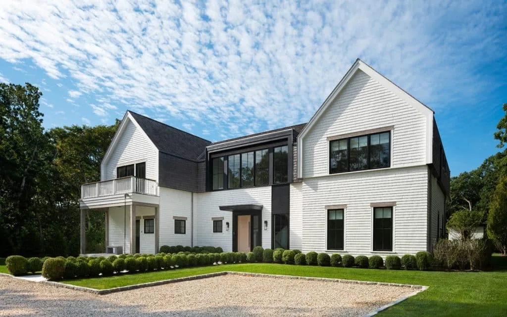 The Amagansett House features a symmetrical facade with white shingles and two chimneys connected by a large windowed section.