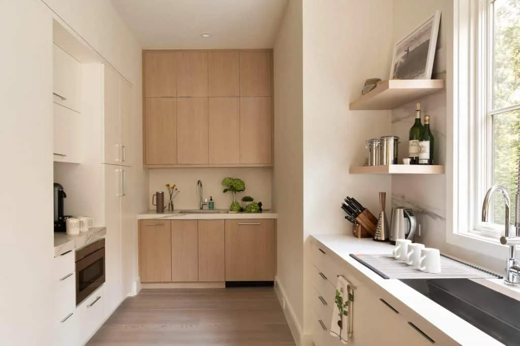 Amagansett House kitchen with vertical wood cabinets, marble backsplash, and two sinks by a large window.