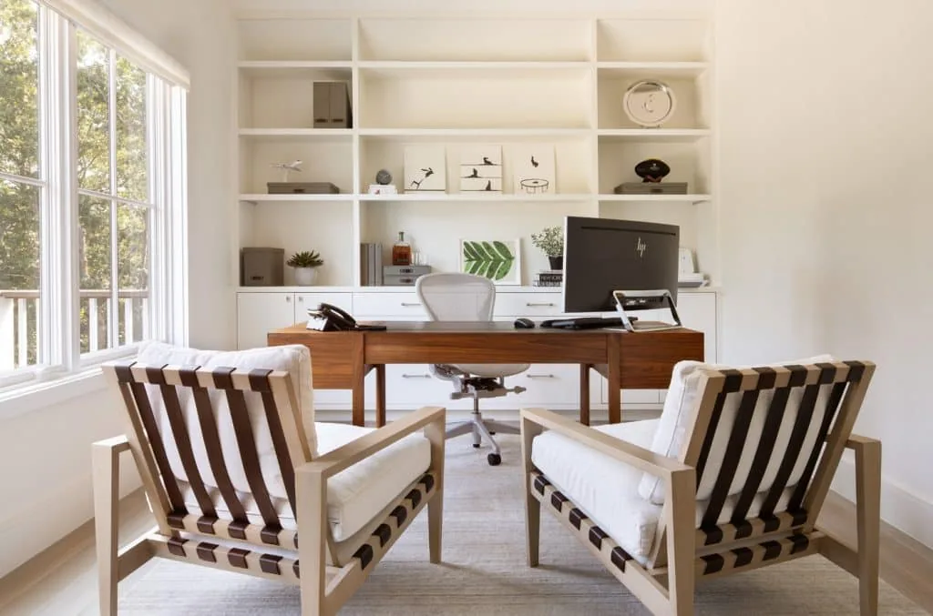 Home office features modern armchairs, a wooden desk, and built-in white shelving with decor.