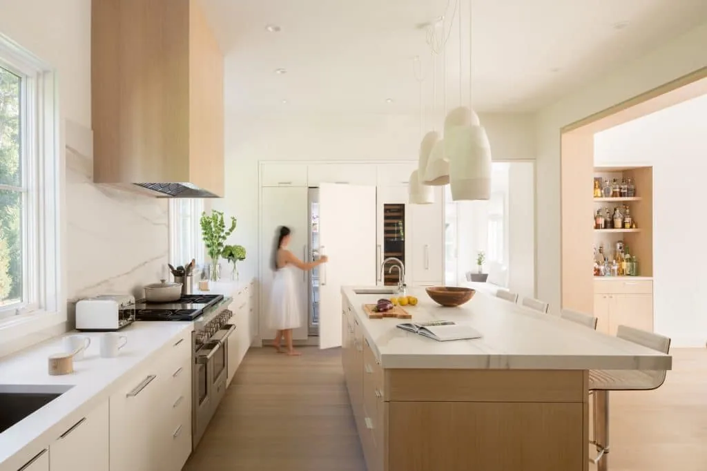 Modern kitchen features a large island with pendant lights, built-in refrigerator, and wood accents by Hacin for the Amagansett House.