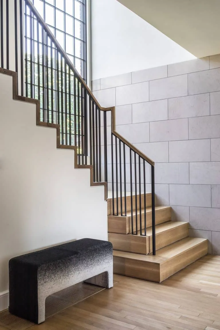 Interior staircase with wood steps and metal railing near a tall window and a small upholstered bench.