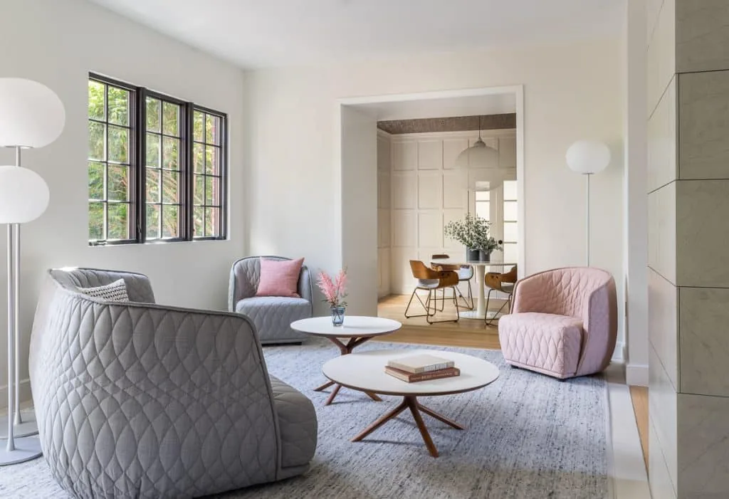Living room featuring quilted armchairs, round wooden coffee tables, and a view into a dining area with a paneled wall and pendant light.
