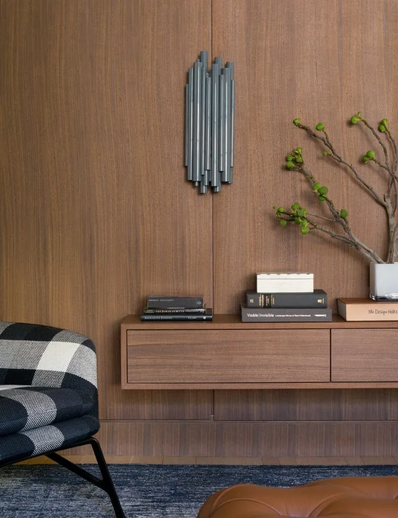 A wood-paneled wall features a floating cabinet with books and a vase, complemented by a modern metal sconce and a plaid upholstered chair.