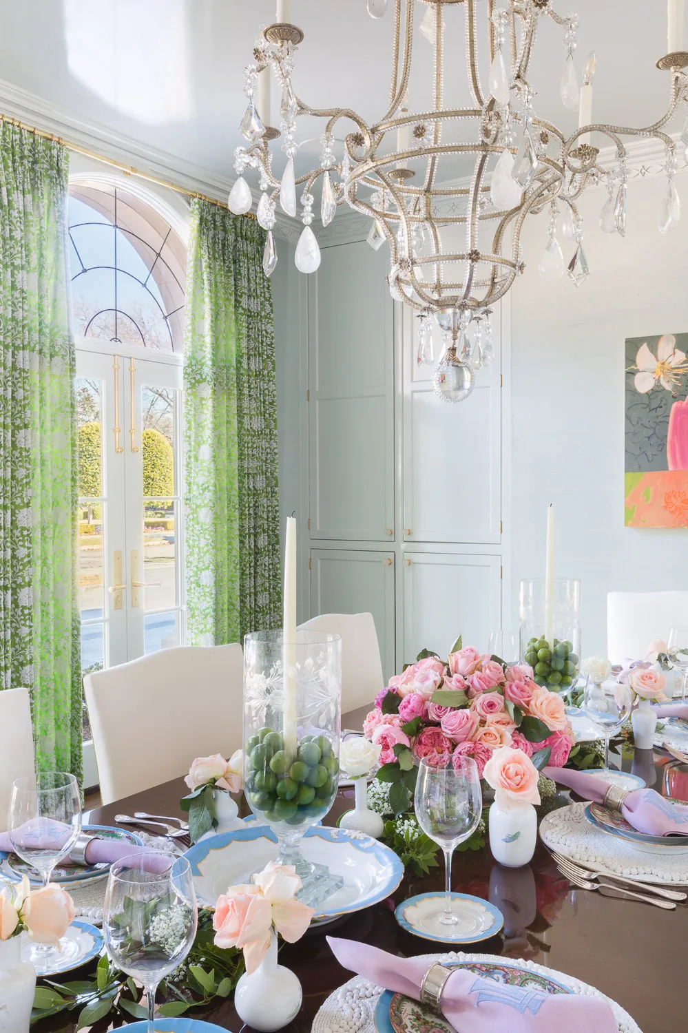 Dining area with floral curtains, elegant chandelier, and table set with flowers and glassware.