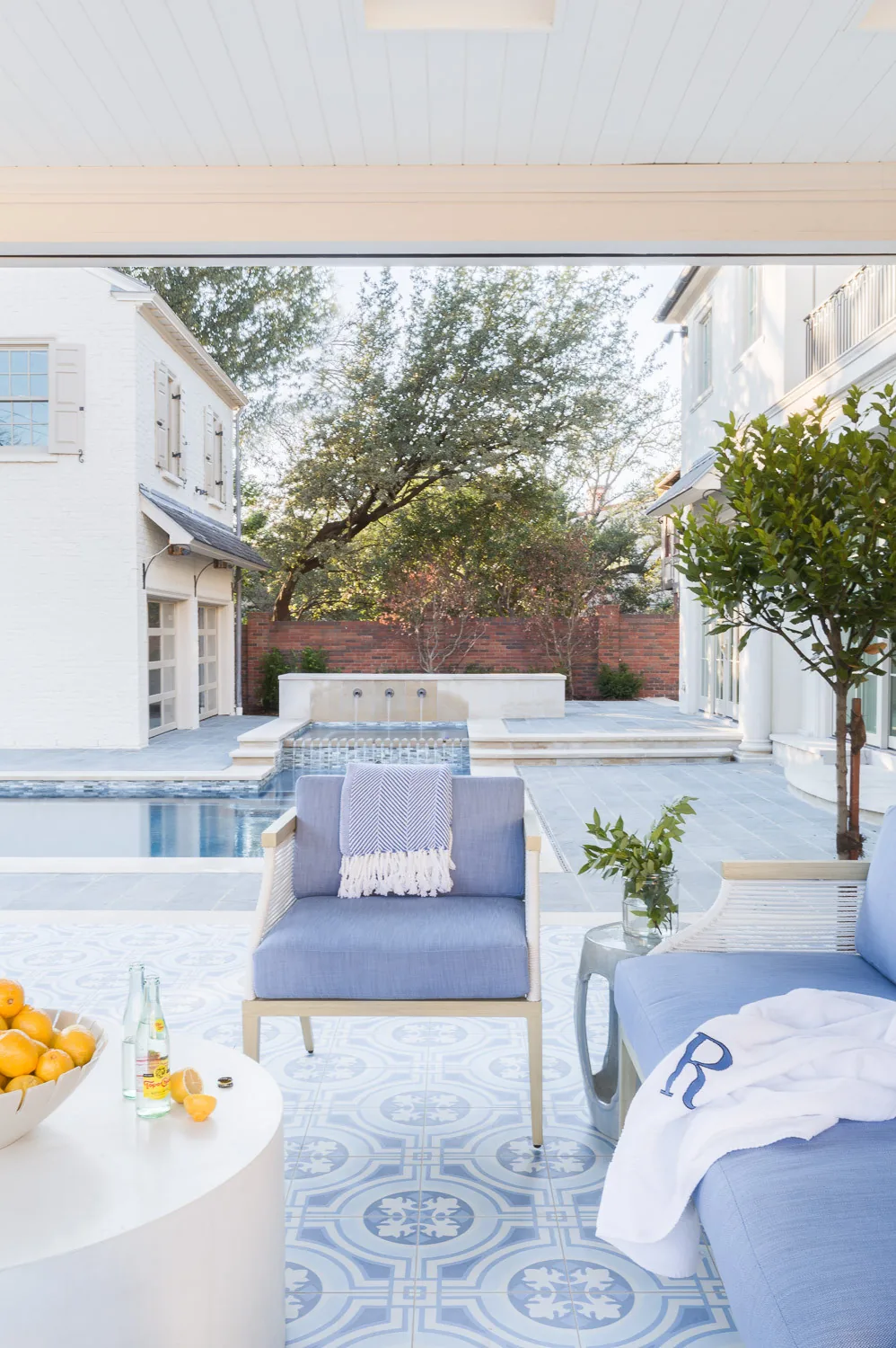 Covered patio with patterned blue and white floor tiles, blue cushioned chairs, and pool with water feature in the background.