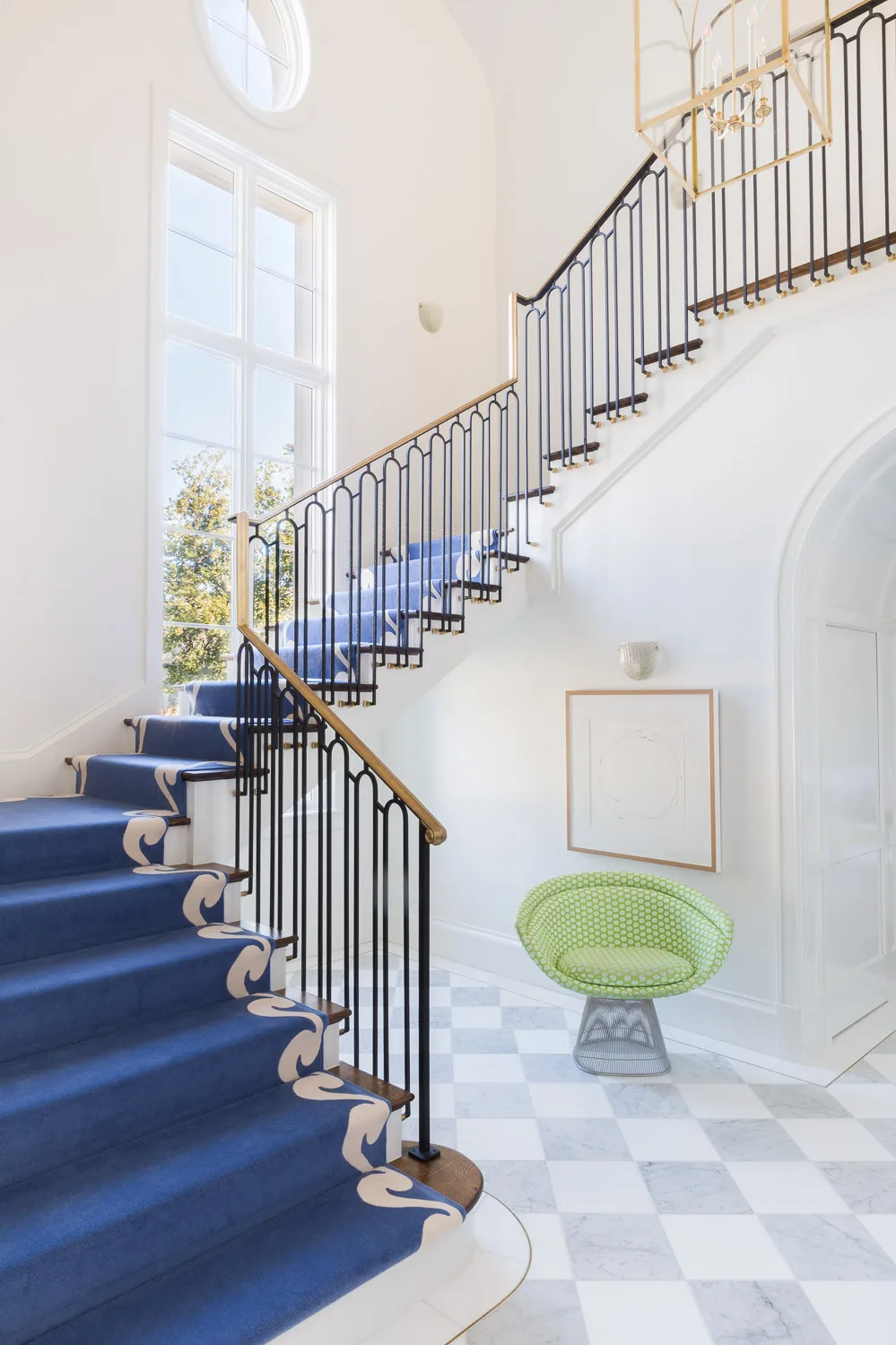 Entry stair with patterned blue carpet, checkerboard floor, and black metal railing with brass handrail lighting an elegant space.