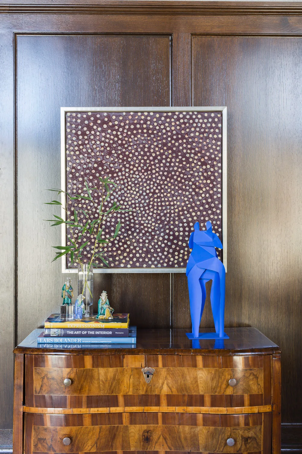 Wood panel wall features framed abstract artwork above a vintage wood chest with books and decorative items.