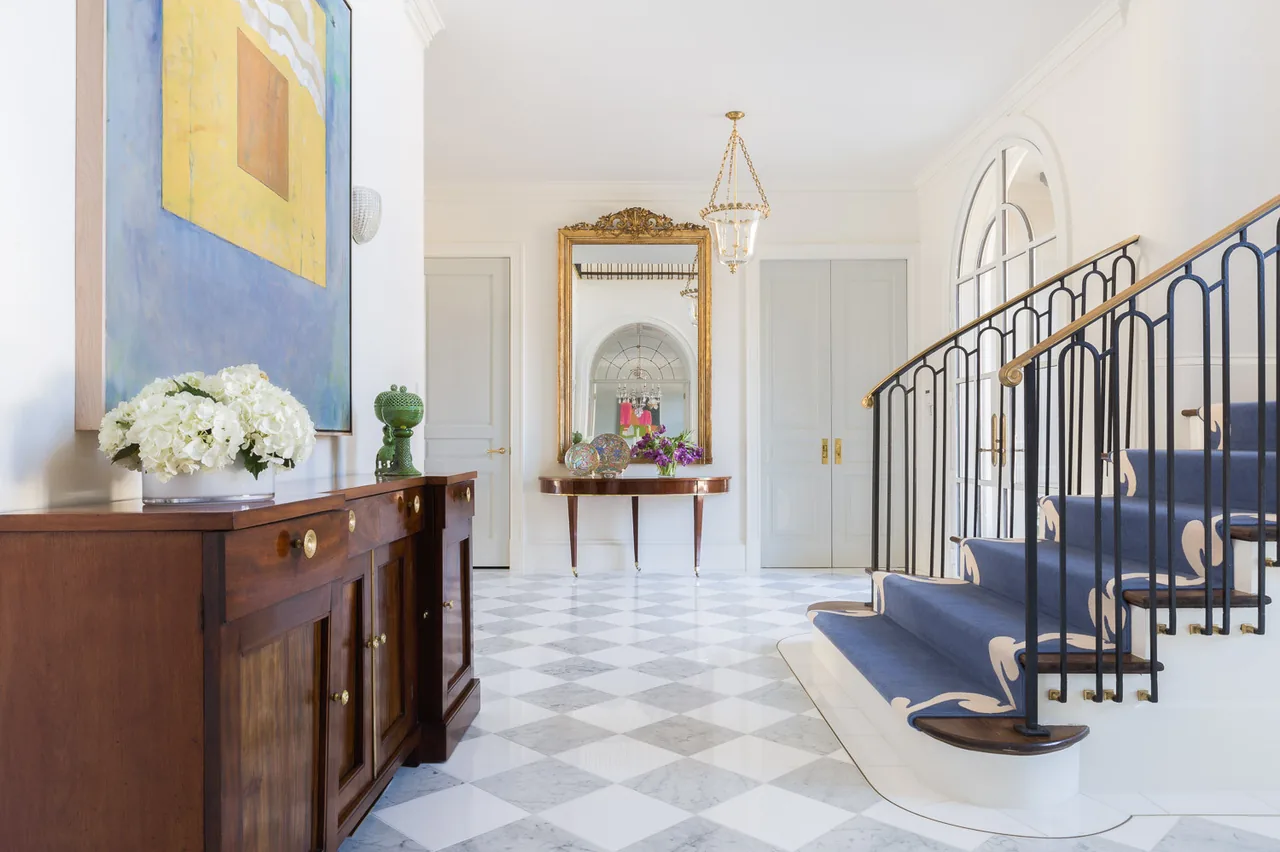 Entry hallway featuring a checkerboard marble floor, a staircase with patterned blue carpet and metal balusters, and a gold-framed mirror on the wall.