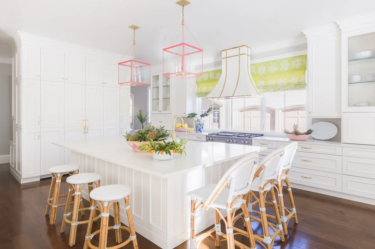 Belclaire kitchen features a large white island with rattan stools, pink pendant lights, and floral green patterned window shades.