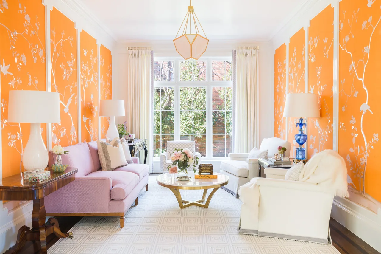 Living room with vivid orange floral wallpaper, pastel pink and white seating, and a round coffee table under a geometric pendant light.
