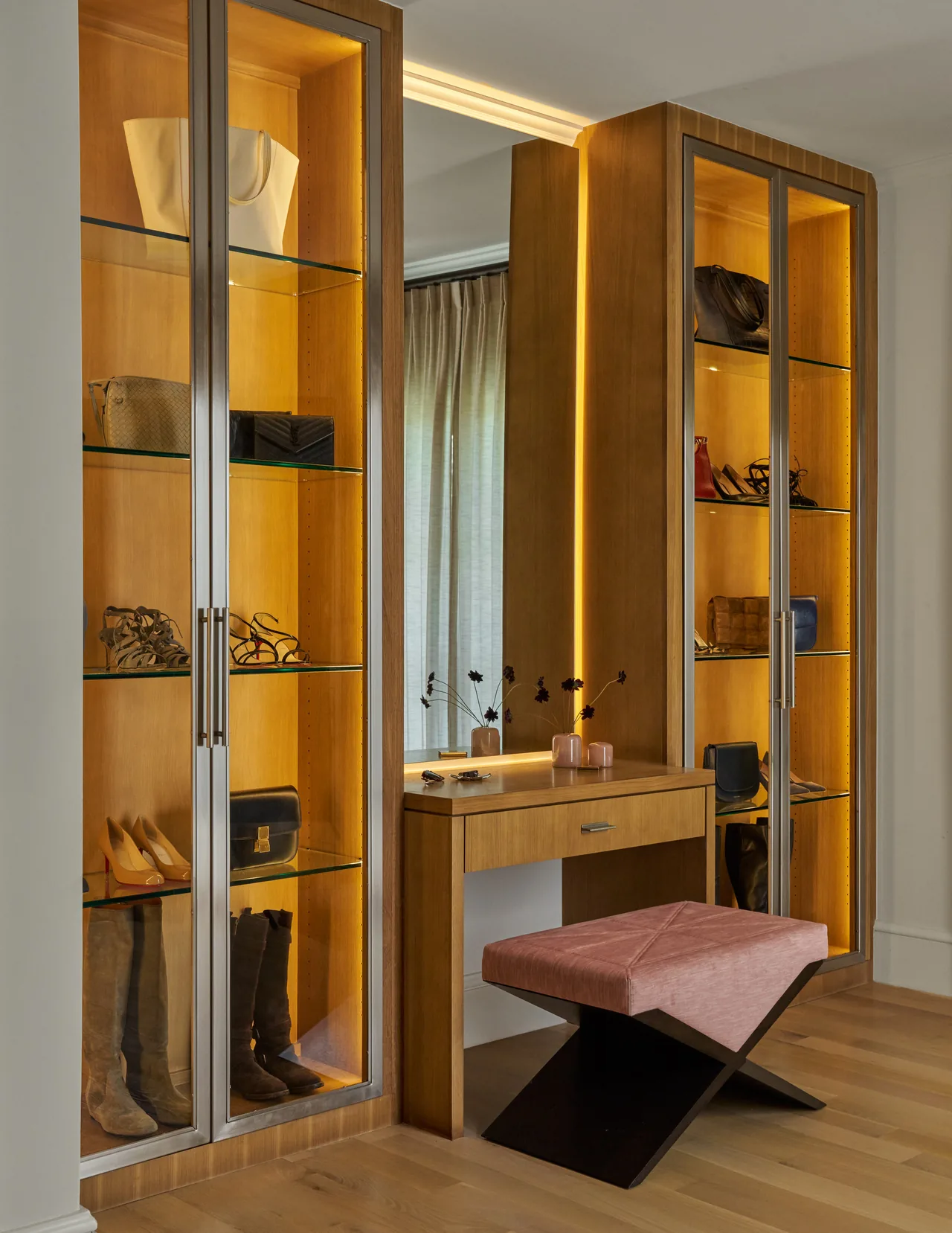 Walk-in closet vanity area with wood cabinetry, glass shelving, and integrated lighting alongside a pink cushioned bench.