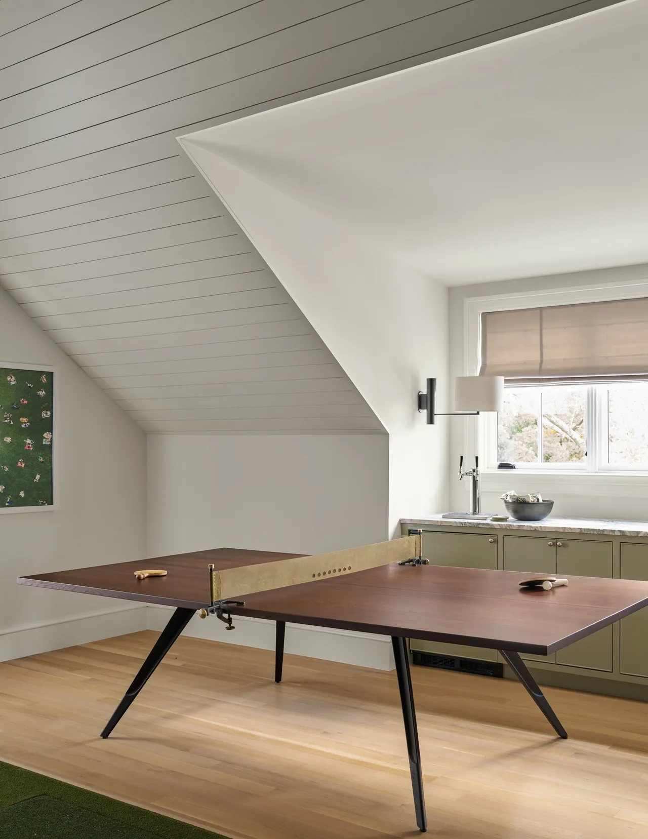 Game room with angled shiplap ceiling, wood ping-pong table, and green lower cabinets beneath a window with a Roman shade.