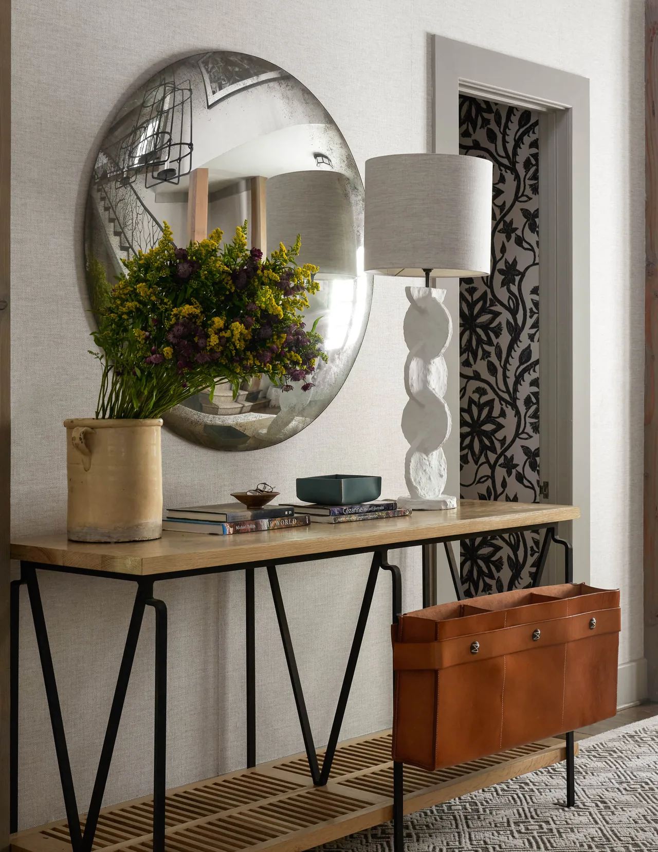 Wooden console table with metal legs and decorative items including a textured lamp and flower vase beneath a circular wall mirror.