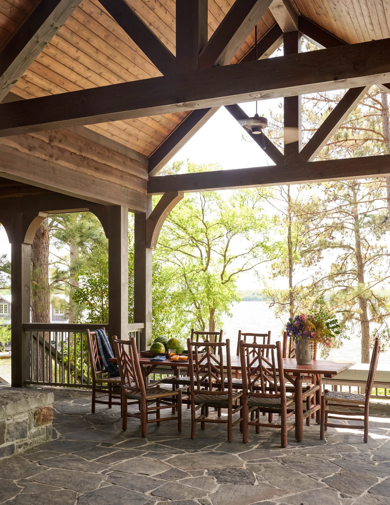 Covered patio dining space with wooden furniture and natural stone floor, overlooking a lakeside view.