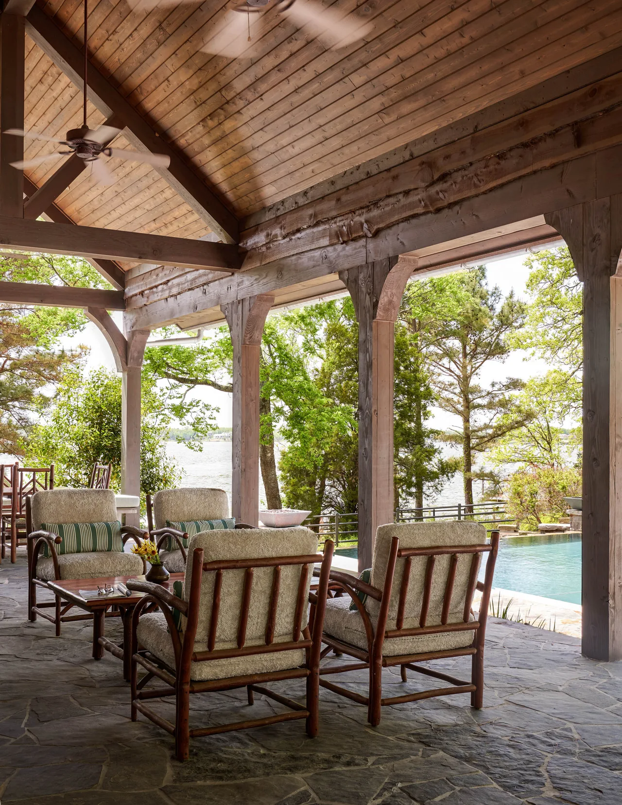 Covered patio seating area with wooden structure and stone floor, looking out towards a pool and lakeside scenery.