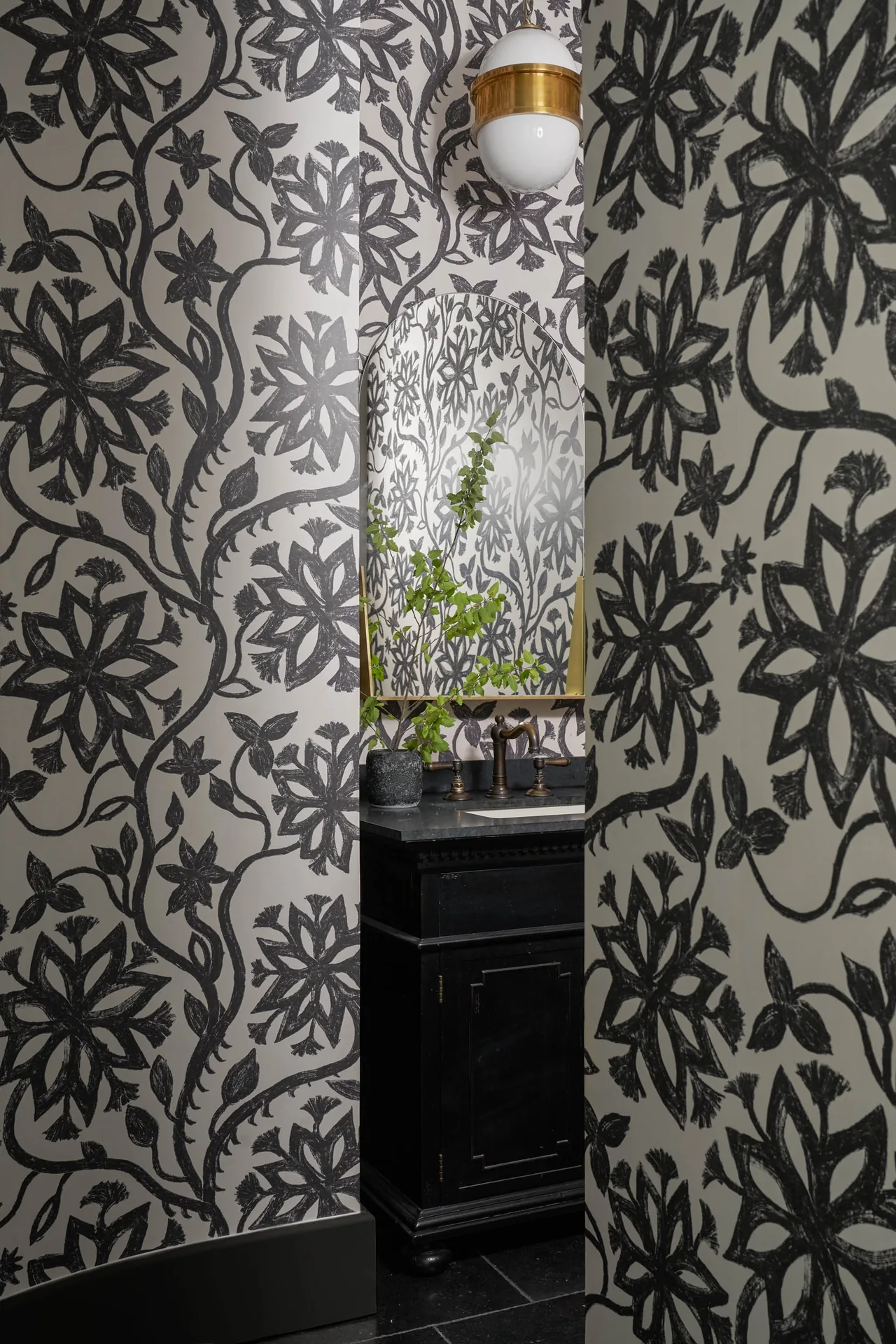 Powder room with black floral wallpaper, black vanity, brass fixtures, and a round mirror reflecting the pattern.