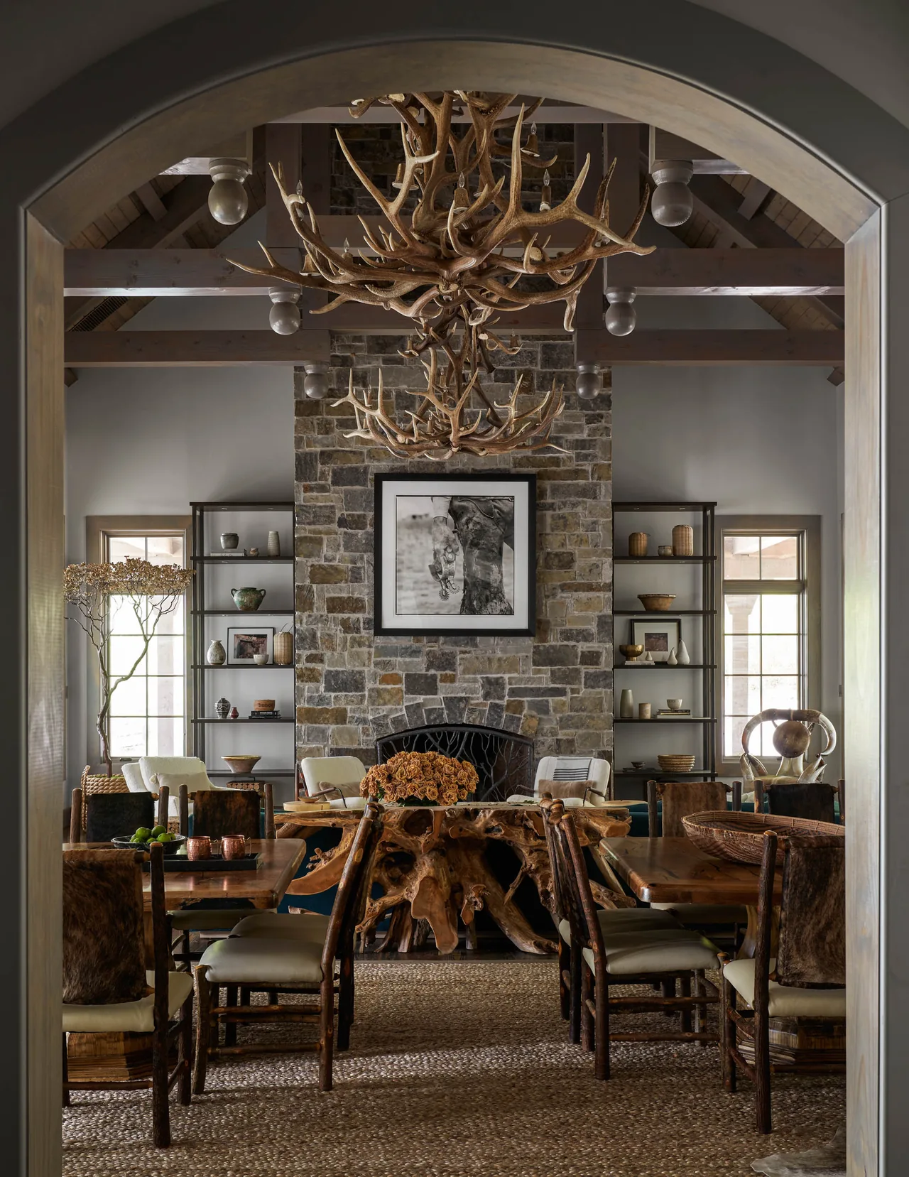 Dining room features a stone fireplace, wooden tables, and twin antler chandeliers under exposed beam vaulted ceiling.