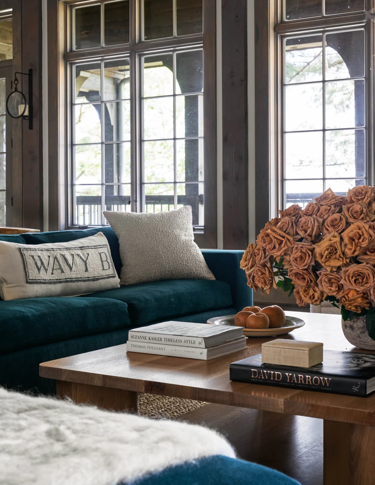 Living room view showing a teal sofa, divided lite windows, and a natural wood coffee table with books and a floral centerpiece.