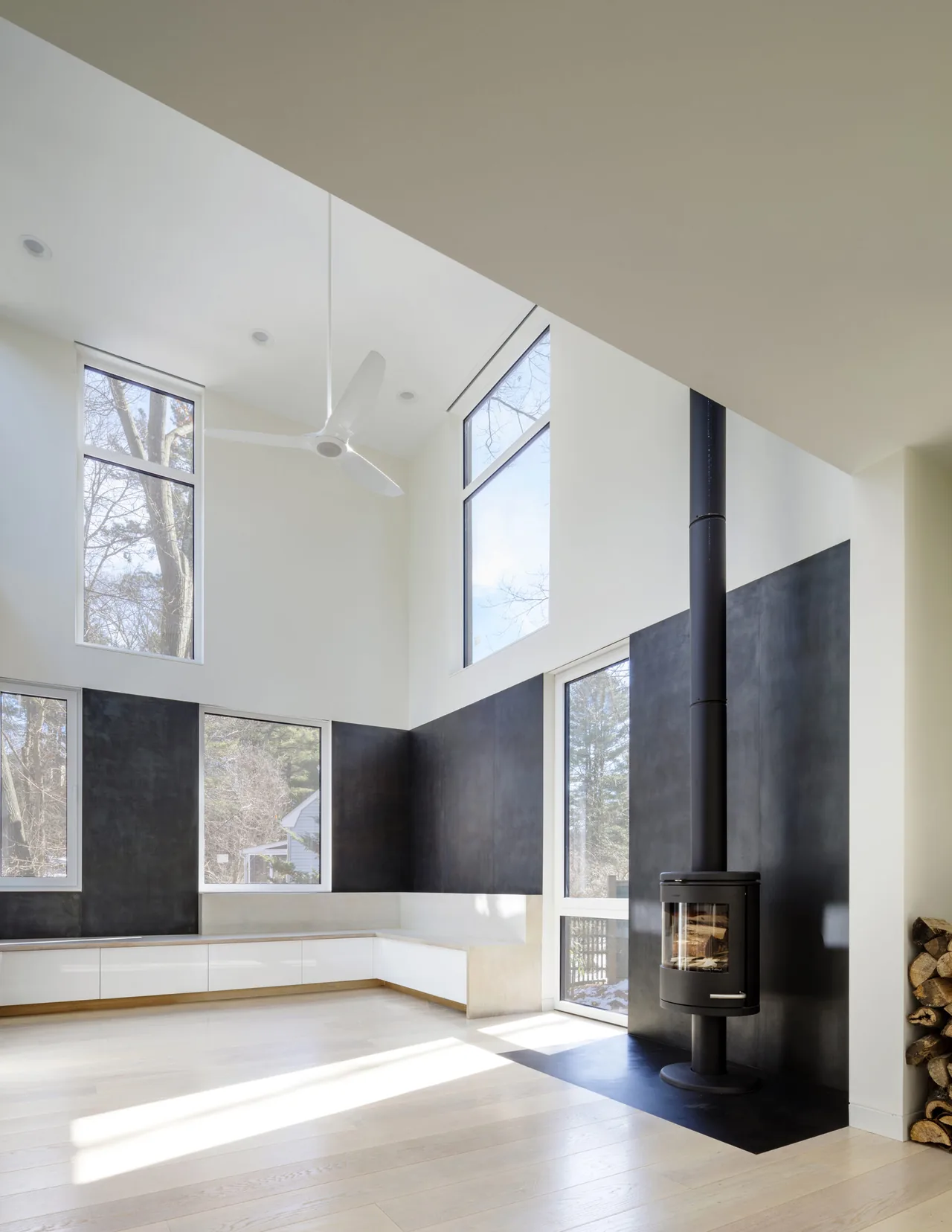 Living room with a double-height ceiling, black wood stove, black paneling, and large windows providing natural light.