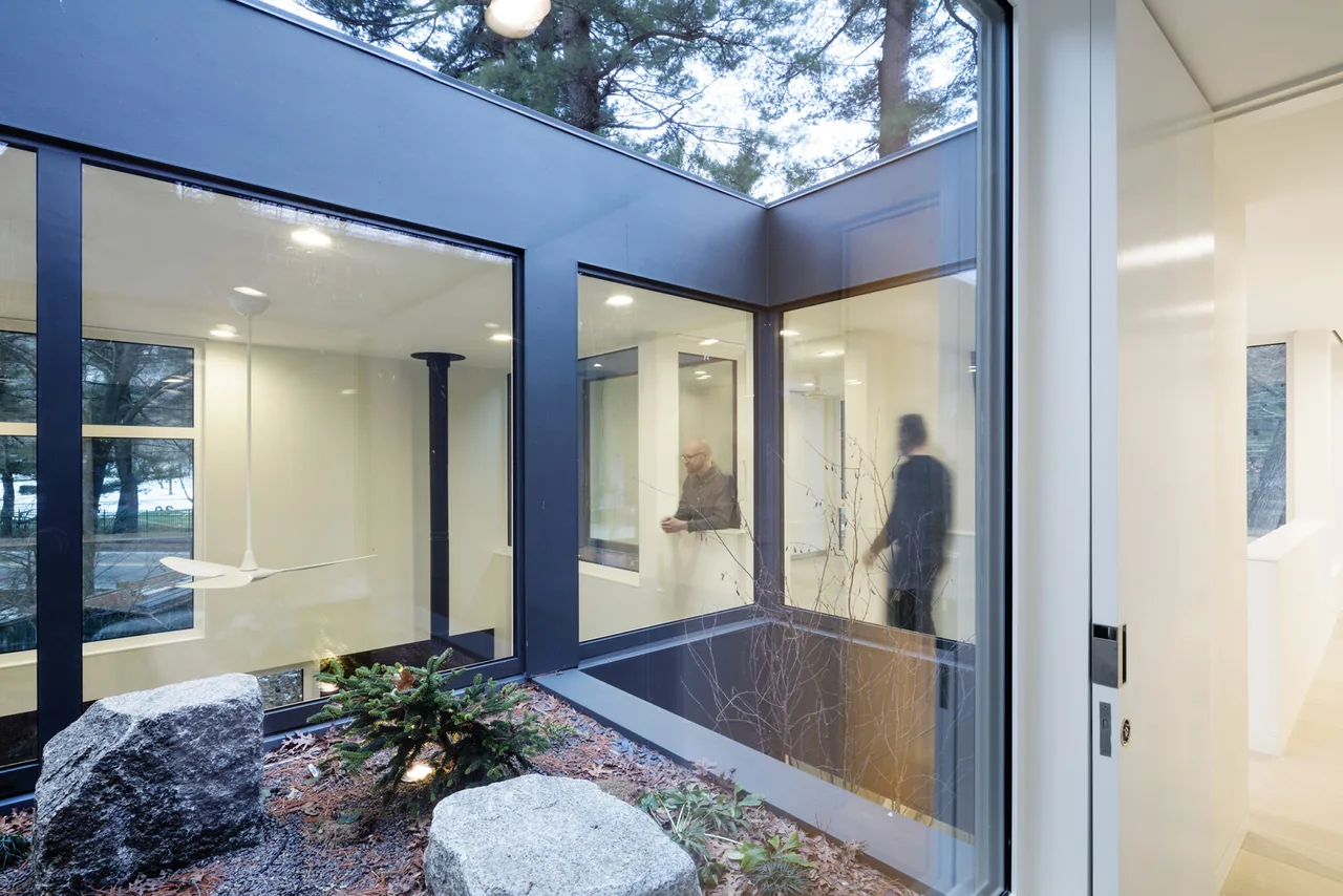 Interior courtyard with stone landscaping framed by large black-framed glass windows showing a double-height room with a black wood stove.