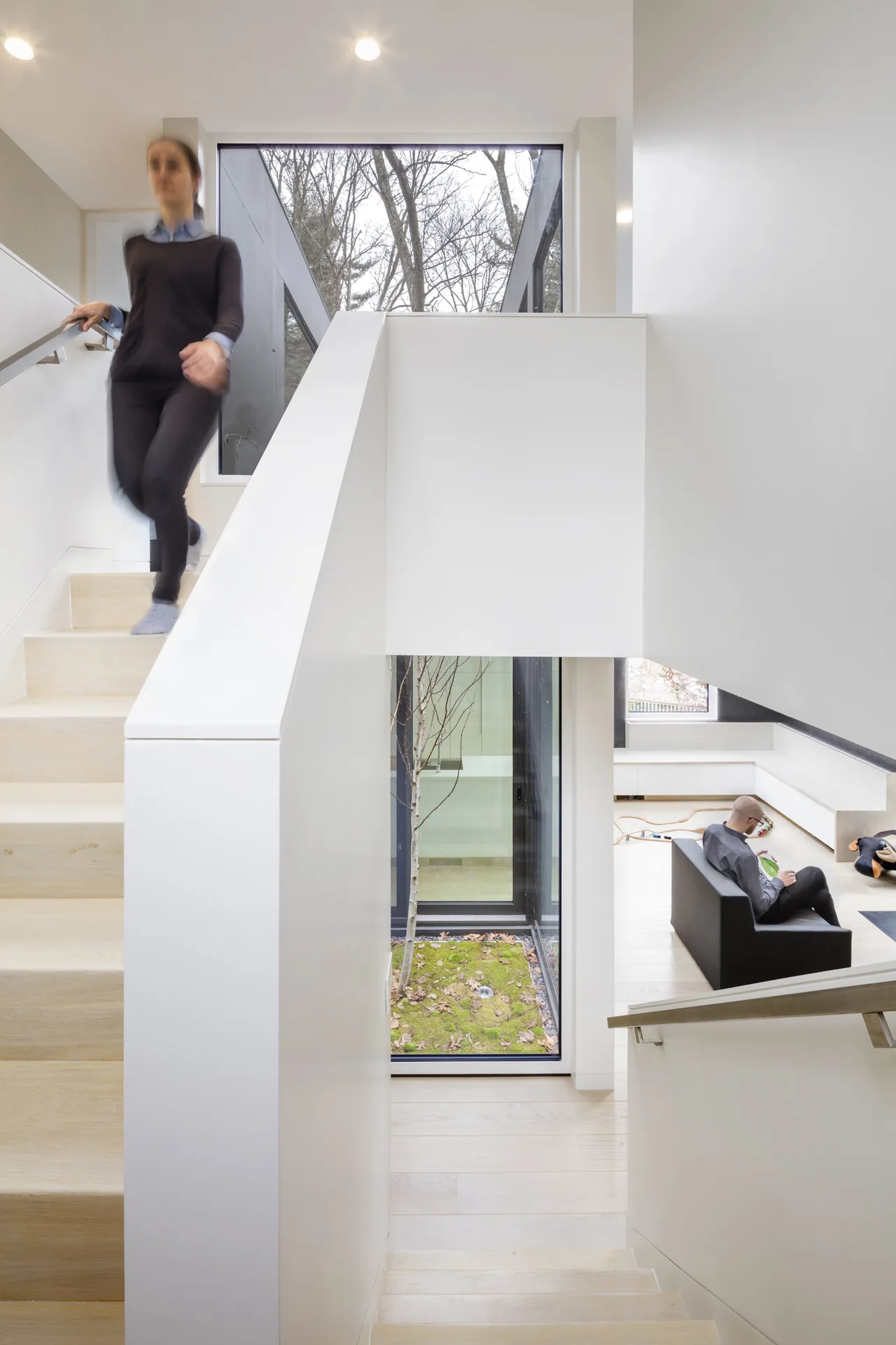 Interior stairwell view showing light wood stairs, a courtyard with a tree, and part of the living room with modern furnishings.