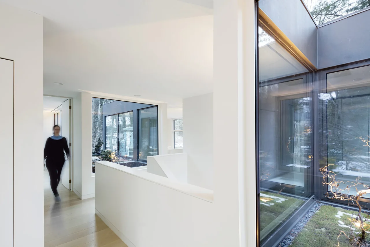 Hallway with light wood floor and double-height ceiling looks out on an interior courtyard with large square windows and black frames.