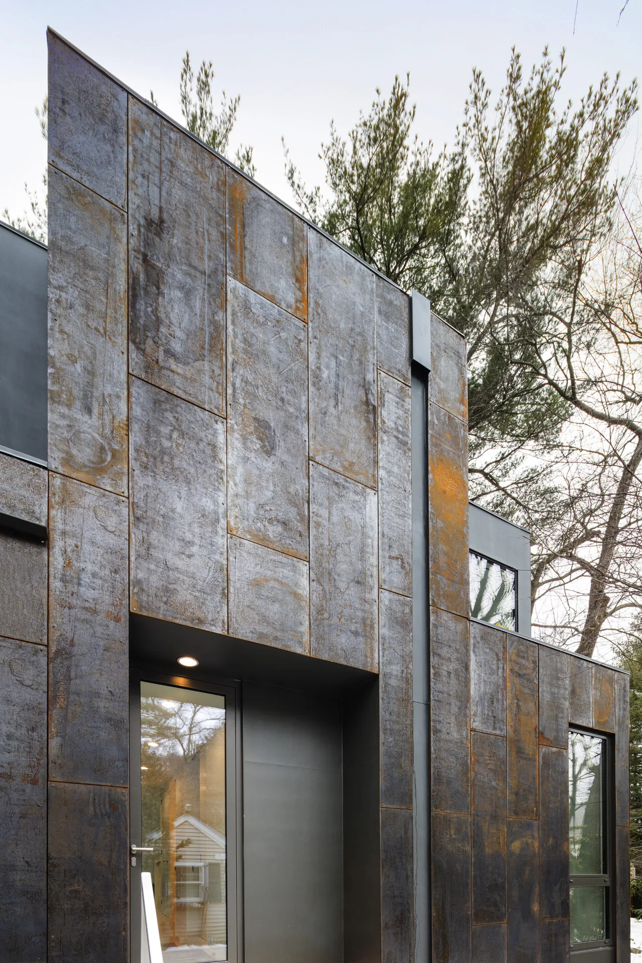 Exterior view of a two-story cube-shaped house clad in rusted metal panels with black-framed windows.