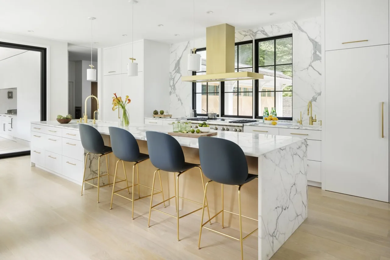 The kitchen features a marble waterfall island, gold fixtures, black window frames, and dark blue bar stools.