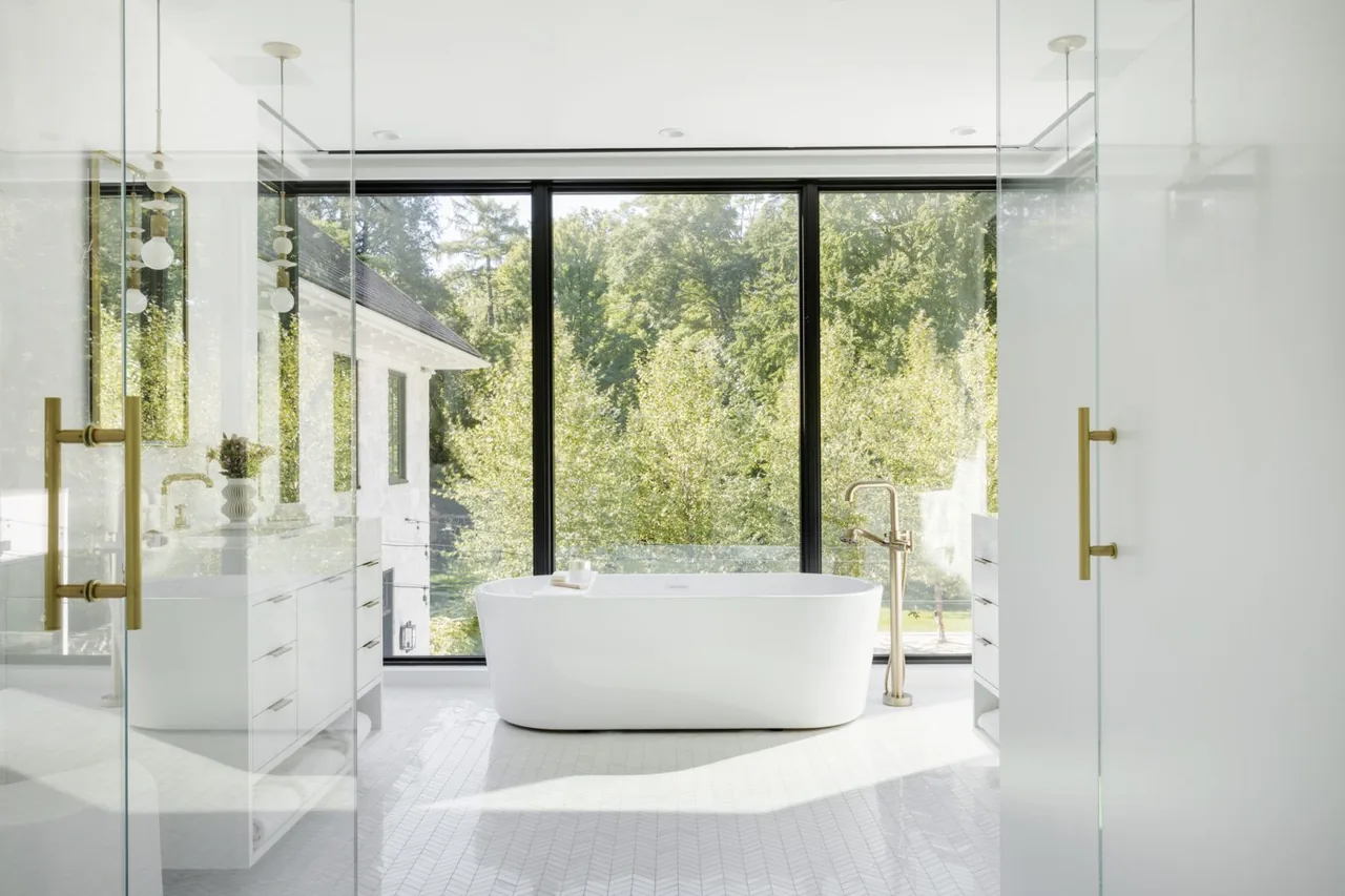 A freestanding tub faces large floor-to-ceiling windows with a lush green outdoor view in a bright bathroom.