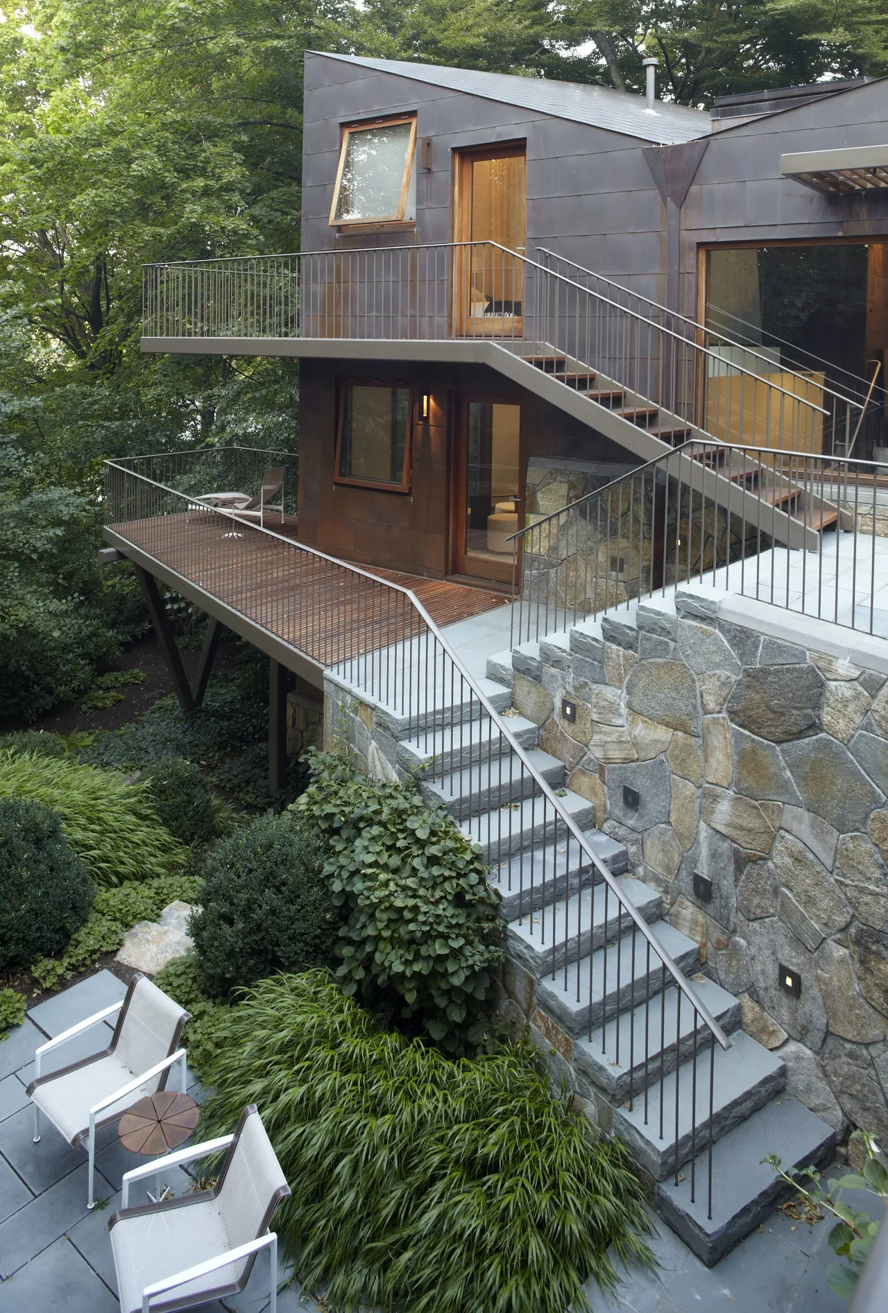 Copper House by Kennard Architects features metal railing stairs and a stone retaining wall on a wooded lot.
