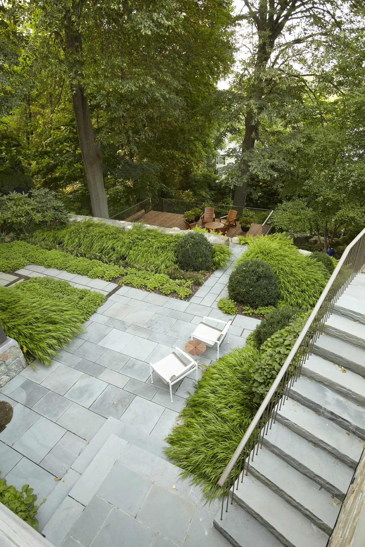 Backyard patio area featuring stone tile paving, stairs with metal railing, and a raised wooden deck with seating beyond a stone retaining wall.