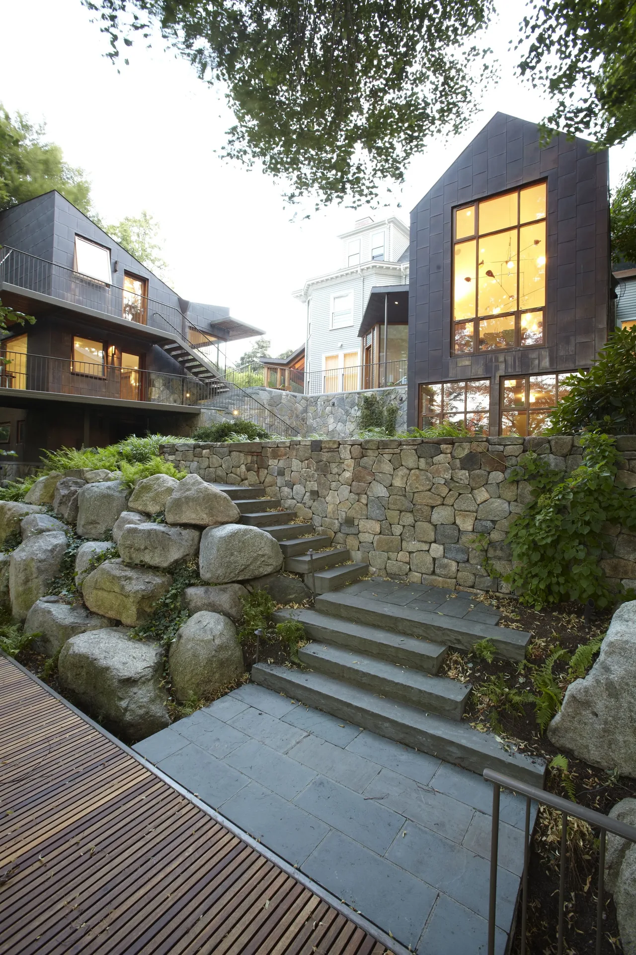 The Copper House in Boston features black metal exterior, stone retaining walls, and extensive large windows overlooking landscaped terraces.