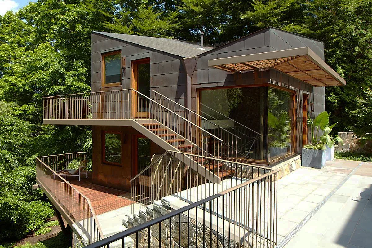 The Copper House by Kennard Architects features large windows, a stone patio, and outdoor metal stairs within a wooded setting.
