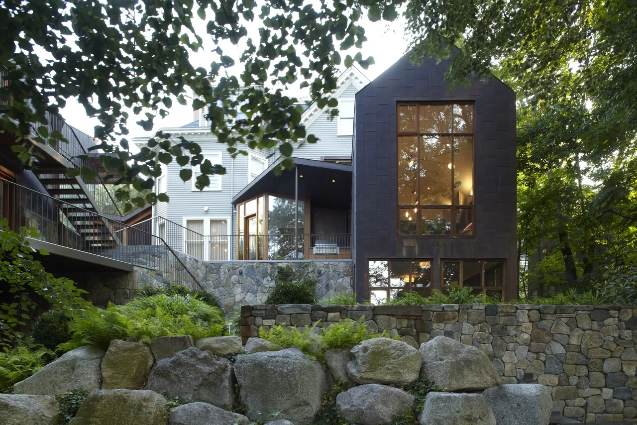 Stone retaining walls and a black modern house extension with large windows surrounded by trees and greenery.