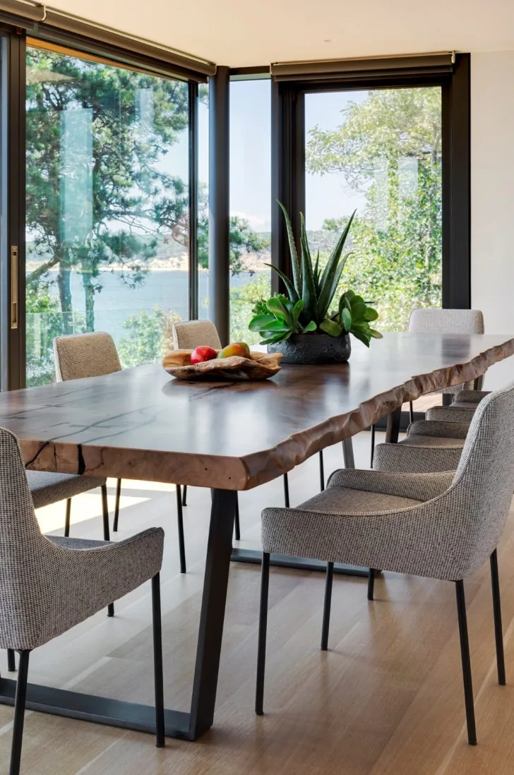 Dining area with a live-edge wood table and gray upholstered chairs framed by large black-windowed corner glazing.