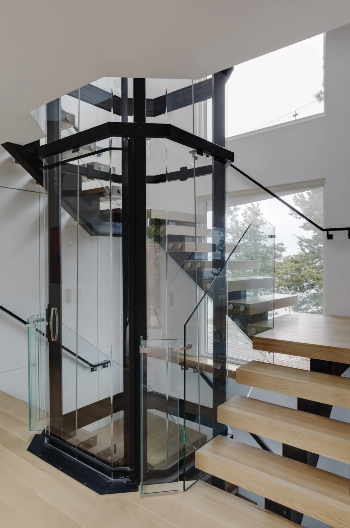 Interior view of a glass elevator shaft integrated with a floating wood stair in a modern home.