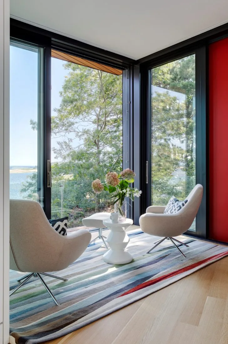 Modern sitting nook with beige armchairs, a white side table, and floor-to-ceiling black-framed glass windows overlooking trees.