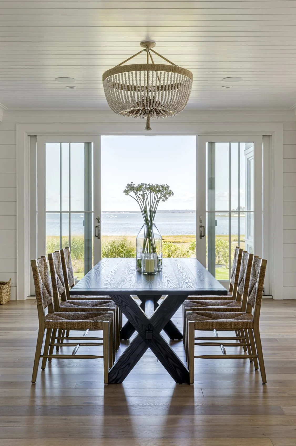 Dining area features an X-base wood table and wicker chairs facing sliding glass doors with a waterfront view.