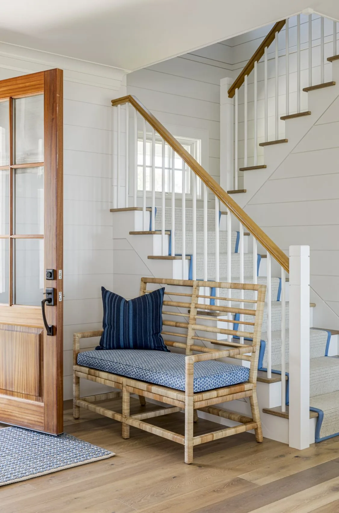 Entry space featuring a wooden door, stair with wood handrail and white spindles, and a natural wood bench accented with blue cushions.