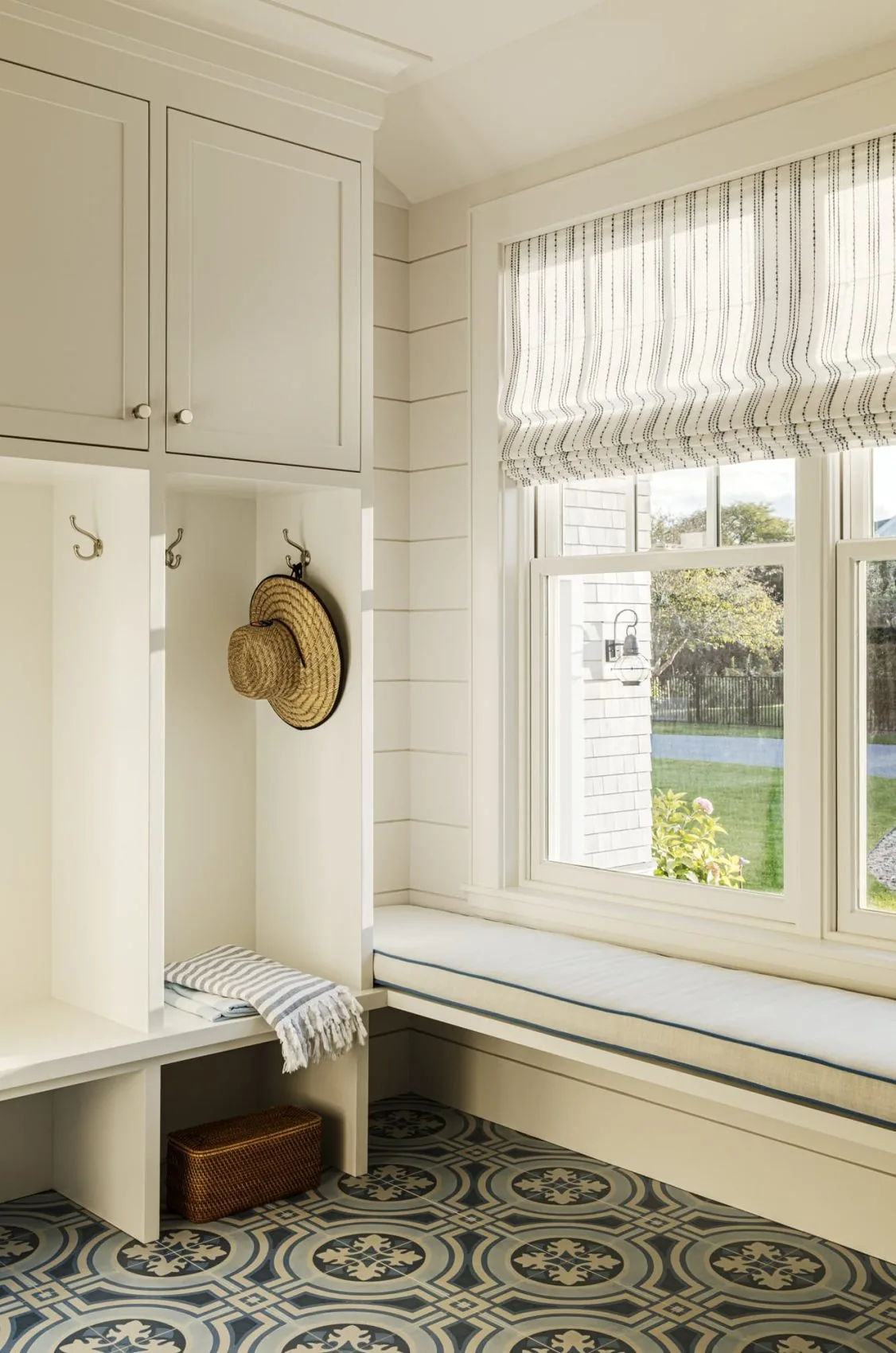 Salten Point mudroom features white built-in storage cubbies with brass hooks and a cushioned bench by a large window.
