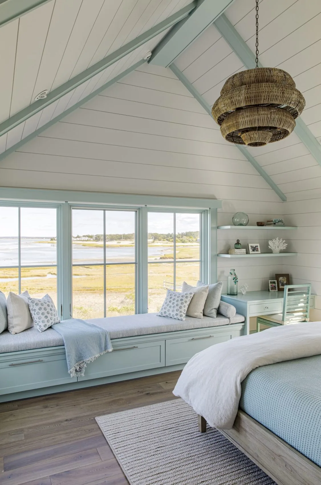 A bedroom features a light blue built-in window seat, vaulted ceiling with exposed beams, and a woven chandelier overlooking a marsh landscape.