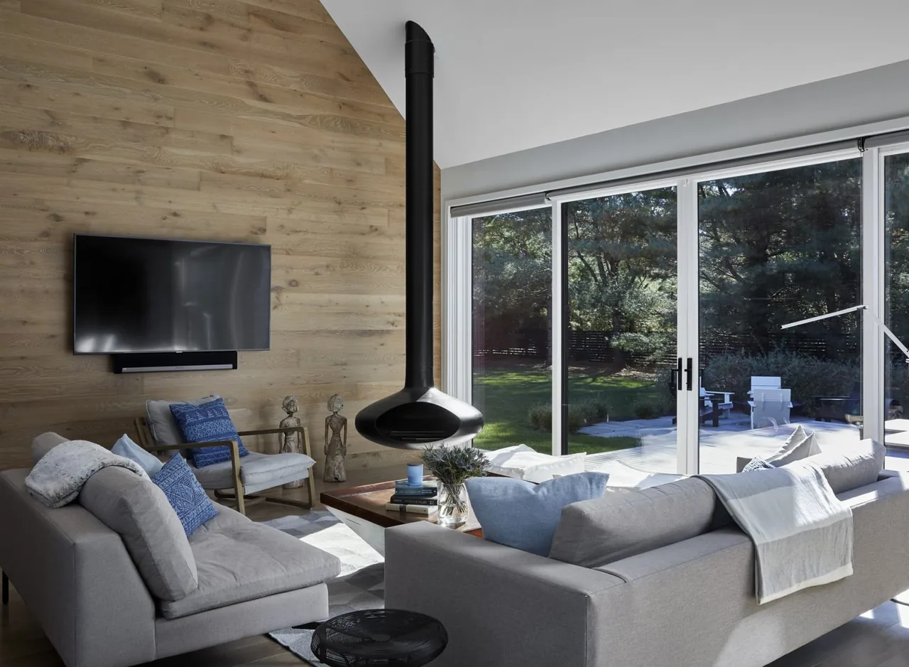 This living room features a wood-paneled wall with wall-mounted TV, central suspended black fireplace, and large sliding glass doors opening to the outdoor area.