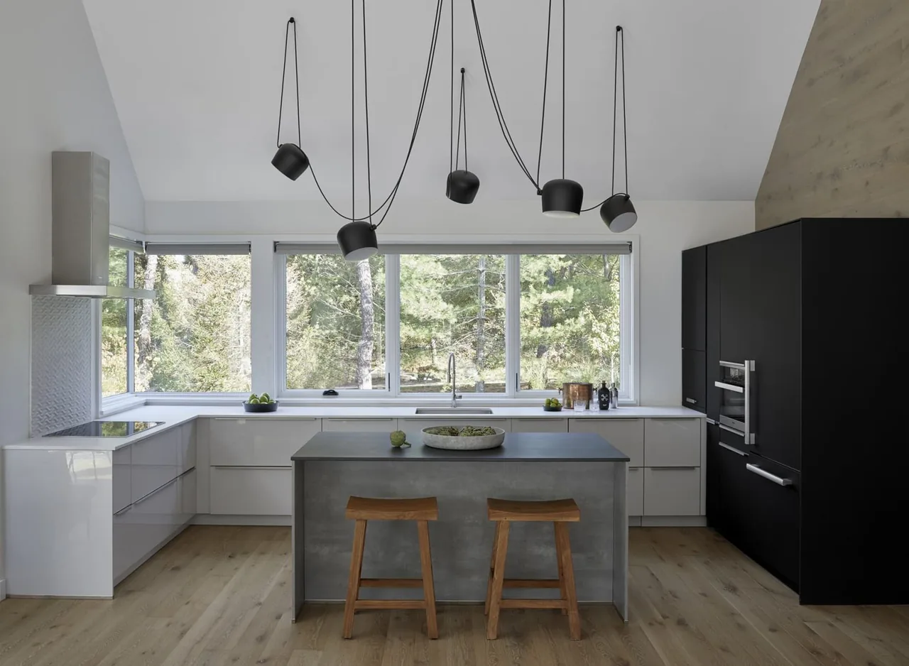 Modern kitchen with a central gray island, white cabinets, black built-in appliances, and large windows providing a forest view.