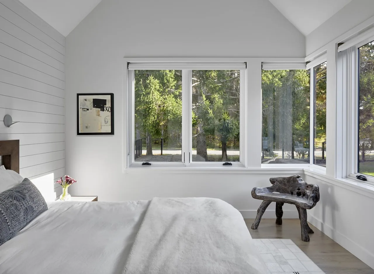 A minimalist bedroom corner featuring white shiplap walls and expansive windows with views of trees outside.