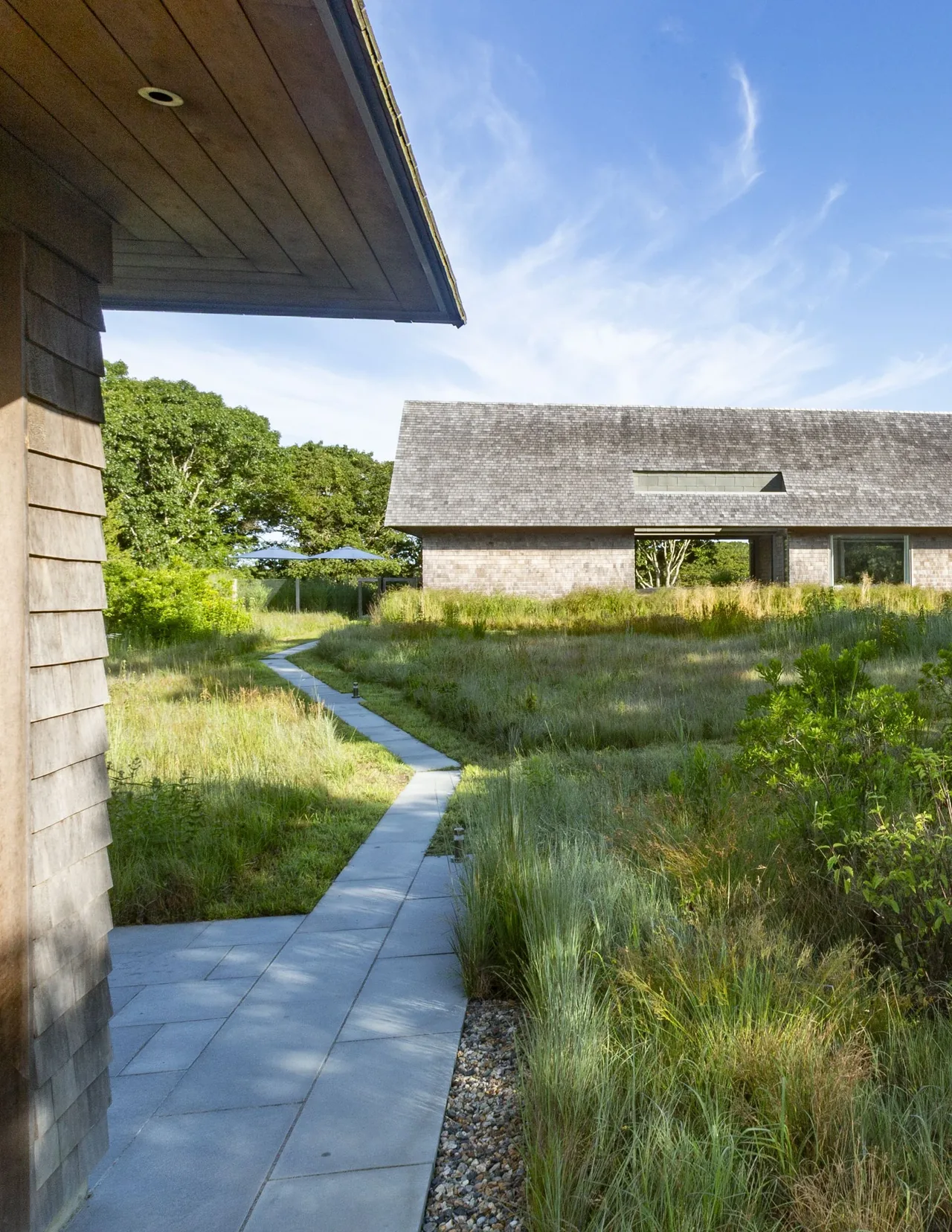 The Wink House features a stone walkway amidst natural landscaping linking two wood-shingled buildings with pitched roofs.