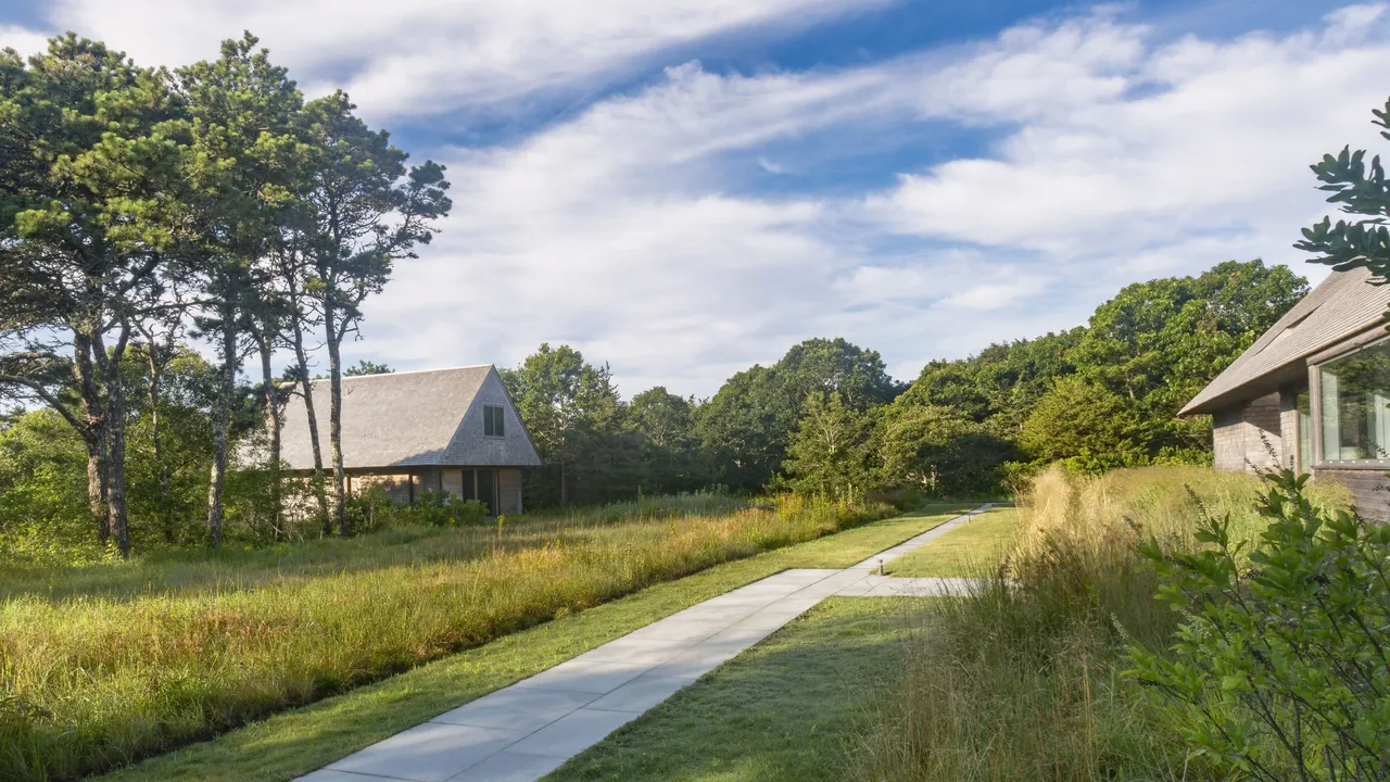 Wink House features natural landscaping with tall grasses and a stone walkway between two pitched-roof buildings.